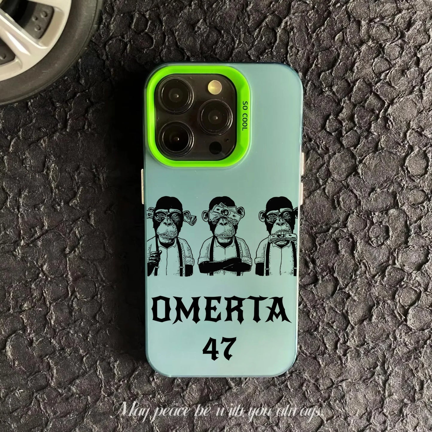 Coque de téléphone verte avec trois singes dessinés et texte OMERTA 47 sur surface asphaltée